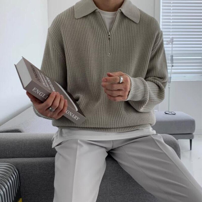 Mann in beigem Strickpullover hält ein englisches Wörterbuch. Stilvolle, moderne Herrenmode, gemütliches Wohnzimmer, minimalistische Einrichtung.