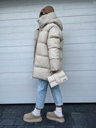 Damen Winterjacke in Creme, gesteppt, mit Kapuze. Kombiniert mit blauer Jeans, beiger Mütze und Tasche. Modetrend, warm, stilvoll.