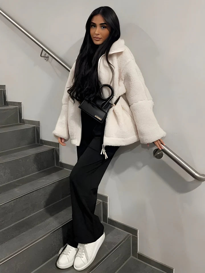 Frau in weißem Teddy-Mantel, schwarzer Hose und weißen Sneakers auf Treppe. Modetrend, Wintermode, stilvolle Damenbekleidung, Streetstyle.