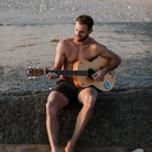 Mann am Strand spielt Gitarre, trägt khakifarbene Badeshorts aus recyceltem Polyester. Sommermode, umweltfreundliche Herrenbekleidung.